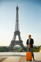 Handsome man woman with orange suitcase standing on Trocadero square. Paris cityscape, view of Eiffel Tower. Travel concept