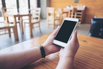 Mockup image of hands holding white mobile phone with blank black screen on wooden table in cafe