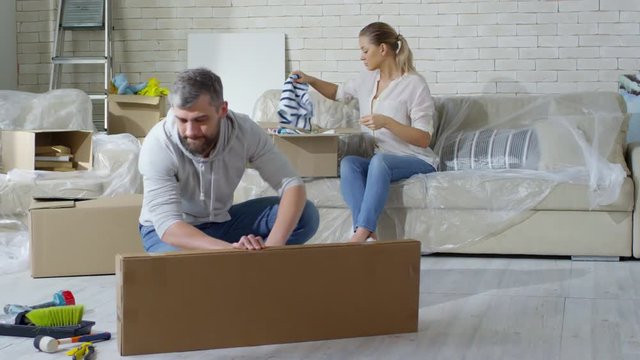 PAN of beautiful woman sitting on sofa covered with plastic wrap and packing children clothes into cardboard box while chatting with bearded husband