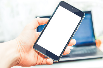 Phone held in hands in the background laptop. A man's hand holds a mobile phone with an empty white display. Businessman working, checking data on the computer, account balance, sales.