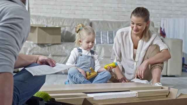 Zoom in shot of cute little girl in denim overalls sitting on floor with cheerful mother and pretending to build disassembled furniture with toy screwdriver while bearded father reading manual