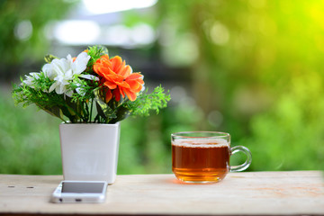 the white and orange  flowers in a white vase with smartphone and a cup of tea  on wooden table and orange light in morning