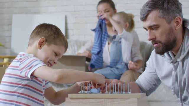 PAN of happy little boy and his bearded father putting wood screws on wooden furniture and arranging them by size while cute girl helping cheerful mother unpacking clothes