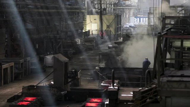 The production process at a steel mill. The crane with hooks moves under the workshop ceiling. Light rays through the dust and air of the workshop. Human labor in Siberia.