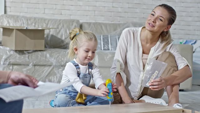 Zoom in shot of adorable girl sitting on floor and pretending to drive screws into disassembled furniture with toy screwdriver while beautiful mother smiling and concentrated father reading manual