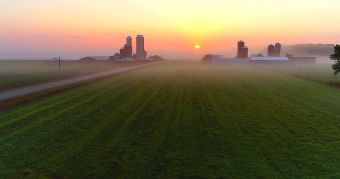 Sunrise over foggy rural agricultural landscape with farms and silos, aerial view.
