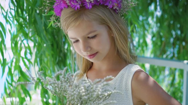 Little blonde with flower's crown posing at camera near tree branches 4K