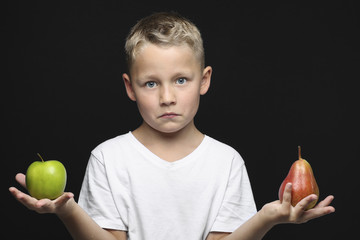 kleiner, blonder Junge hält zweifelnd einen Apfel und eine Birne in der Hand, vor schwarzem...