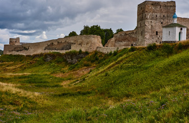 Fototapeta premium Fortress wall on the hill/On the hill there is an ancient fortress. The wall is partially destroyed. Near the square watchtower is a white Orthodox chapel. Russia, Pskov region, landscape