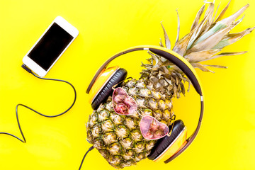 Stylish pineapple in pink glasses listen to music on the smartphone on yellow background top view