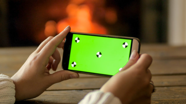 Closeup shot of young woman holding smartphone with empty green screen at burning fireplace. Place for your image or design