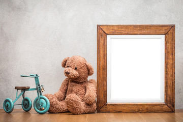 Retro oak wooden photo frame blank and plush Teddy Bear with toy tricycle on the desk front old textured concrete wall background. Vintage style filtered photography