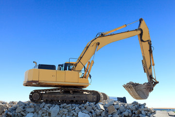 Large backhoe on top of a mound of rocks on a job site. Blue sky is in the background.