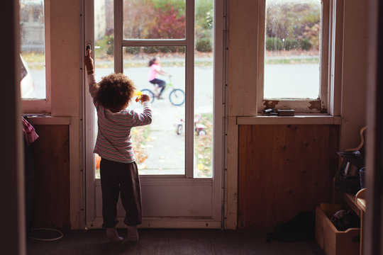 Toddler girl standing in a darkened room looking outward through a window at a girl riding a bicycle