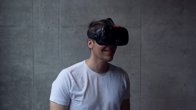 An adult male uses a virtual reality headset, standing behind a grey concrete wall. Medium shot.