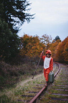 Child in fox costume walking with suitcase along train tracks