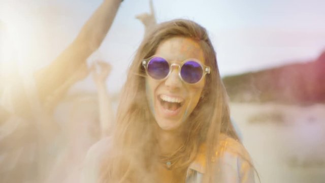 Close-up Portrait of a Beautiful Young Girl with Sunglasses Standing in the Crowd of People Celebrating Holi Festival. People Throwing Colorful Powder in Her Back. 