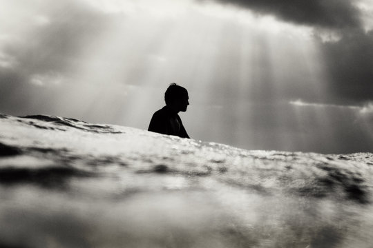 A silhouette of surfer waiting in the lineup with sun rays behind him