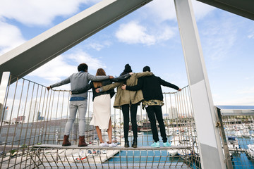 Back view of group of friends embraced looking seaport from bridge.