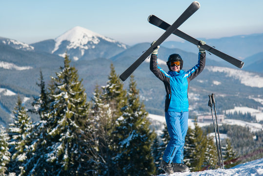 Happy woman skier holding her skis over head, wearing blue ski suit and black helmet standing on top of the mountain on a sunny winter day copyspace skiing happiness active people lifestyle