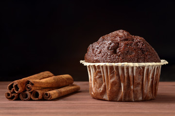 Close up of a delicious chocolate muffin with cinnamon sticks on a wooden table against black background. Space for text