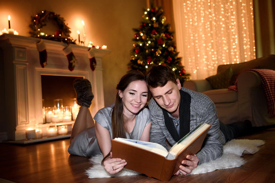 A young family spending evening together looking through some pictures in a big photo album. Couple hugging in front of fireplace at home, reading book.