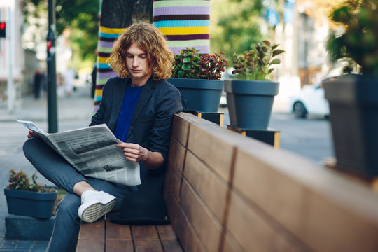 Red haired hipster man sitting on bench reading newspaper