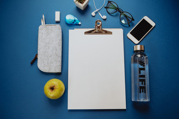 Healthy working concept: a sheet of paper, pencils, water, apple, phone, glasses on a blue background. Top view, flat lay