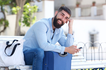 Happy man sitting outside with suitcase and mobile phone