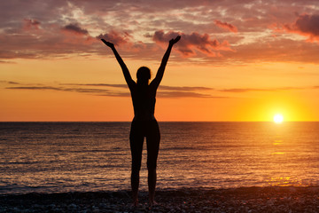 Fototapeta premium Girl standing hands up on the beach at sunset. Silhouette