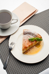 Apple cake with sea buckthorn sauce, icing sugar and basil on white plate. With coffee cup and newspaper.