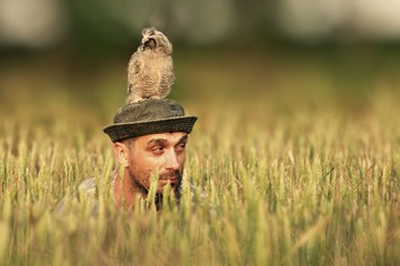 A man looks out of the grass with an owl on his head and looks in different directions.