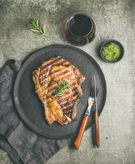Flat-lay of Grilled hot rib-eye beef steak on bone with chimichurri green sauce on black slate round plate and red wine over grey concrete background, top view. Meat high protein dinner concept