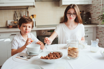 Obraz na płótnie Canvas kid brother and teen sister having breakfast at home in modern white kitchen and eating snacks. Casual lifestyle in real life interior