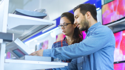 Beautiful Young Couple in the Electronics Store Browsing, Looking for Newest Gadgets, Tablets and Photo/ Video Cameras Presented on the Shelves.