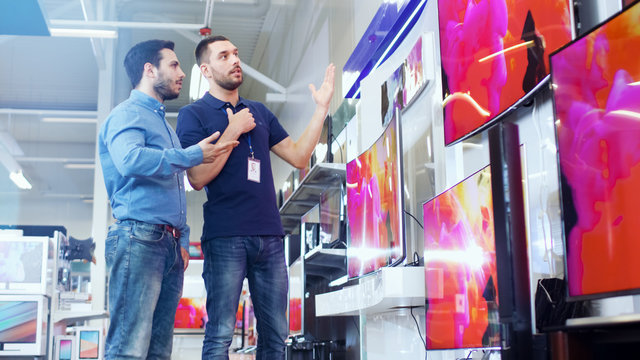 In the Electronics Store Professional Consultant Shows Latest UHD TV's to a Young Man, They Talk about Specifications and What Model is Best for Young Man's Home. Store is Bright, Modern.