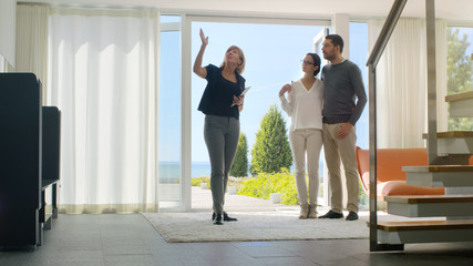 Professional Real Estate Agent Shows Stylish Modern House to a Beautiful Young Couple Who are in the Market for Purchasing/ Renting New Home. House Has Floor to Ceiling Windows and Seaside View.