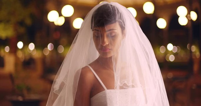 Portrait of elegant bride posing outdoors in evening looking down with veil covering face softly. Lovely black female wearing white veil standing in urban setting with bokeh lights 