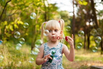 Blonde little girl blows soap bubbles in park, with copy space