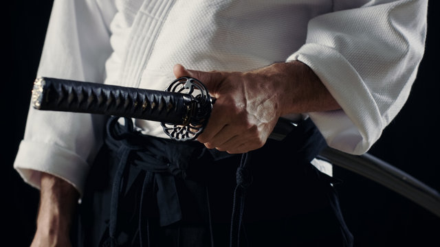 Aikido Master Wearing Traditional Samurai Hakama Clothes Holds His Japanese Sword in Scabbard. He's in the Spotlight Darkness Surrounds Him. Shot Isolated on Black Background.