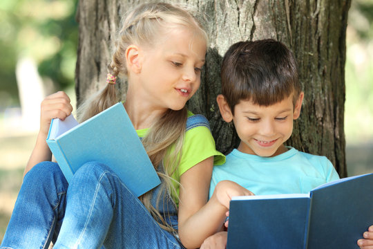 Cute Little Children Reading Books Near Tree In Park