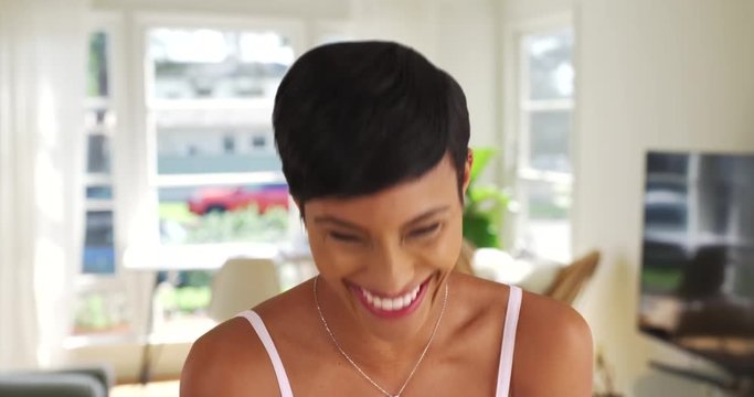 Lovely black female in her 20s laughing at camera joyfully. Young African American woman with cute pixie haircut laughing inside living room 