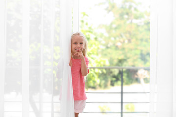 Fototapeta premium Cute little girl hiding behind curtain at home