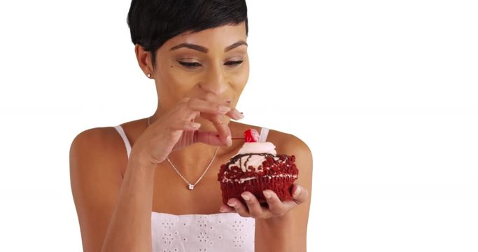 Close up of attractive black female enjoying treat, tasting cupcake frosting in studio with copyspace. Portrait of woman in her 20s savoring big red velvet cupcake, sucking on maraschino cherry 