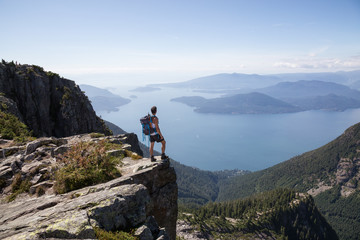 Fototapeta na wymiar Fit and Adventurous Latin American man is hiking on top of a mountain ridge with a beautiful ocean view in the background. Taken at Lions Peaks, North of Vancouver, British Columbia, Canada. 