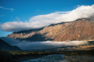 Different double layers of clouds over Stepantsminda Kazbegi village and peak of brown mountain, Georgia. Scenic autumn landscape.