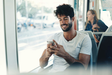Bearded hipster guy is reading emails on a display of a smartphone connected to public wi-fi while sitting in a city bus. Handsome male is looking at the screen of a mobile phone while texting. - obrazy, fototapety, plakaty
