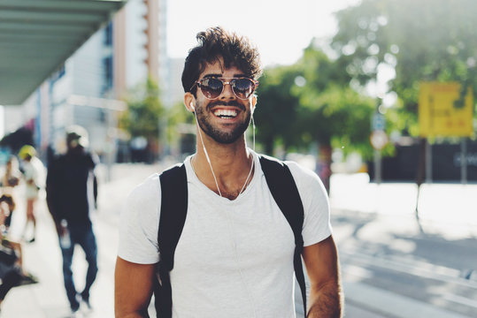 Young travel blogger is ready for a new weekend trip. Bearded backpacker is standing on a blurred station background. Student is relaxing on a weekend. Hipster guy wearing sunglasses smiling at camera