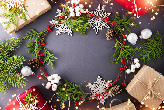 Frame with christmas wreath, gift boxes, branches and snowflakes on dark background top view. Merry christmas greeting card. Winter xmas holiday theme. Happy New Year. Flat lay. Sparkle bokeh lights.