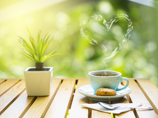 heart smoke on a cup of coffee, cookies, spoon,tissue paper and small tree pot on wooden table in garden with sun light in refreshing morning.green bokeh bush tree background.